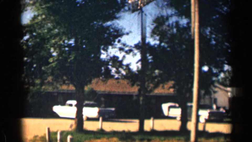 1963 Colorado home-movie frame of a motel lot with trees, parked cars, a white trailer, utility poles, and sunlit ground.