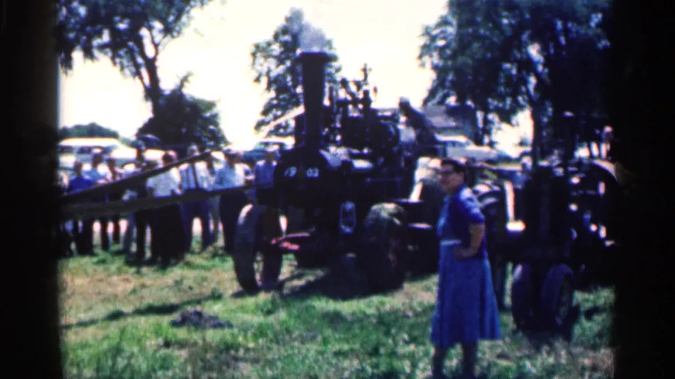 A black steam traction engine is framed with spectators in the background and a woman in a blue dress standing near the machinery.