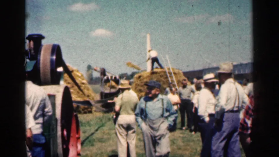 Men and visitors stand near hay handling activity and farm machinery in the 1963 Montgomery, Minnesota outdoor gathering.