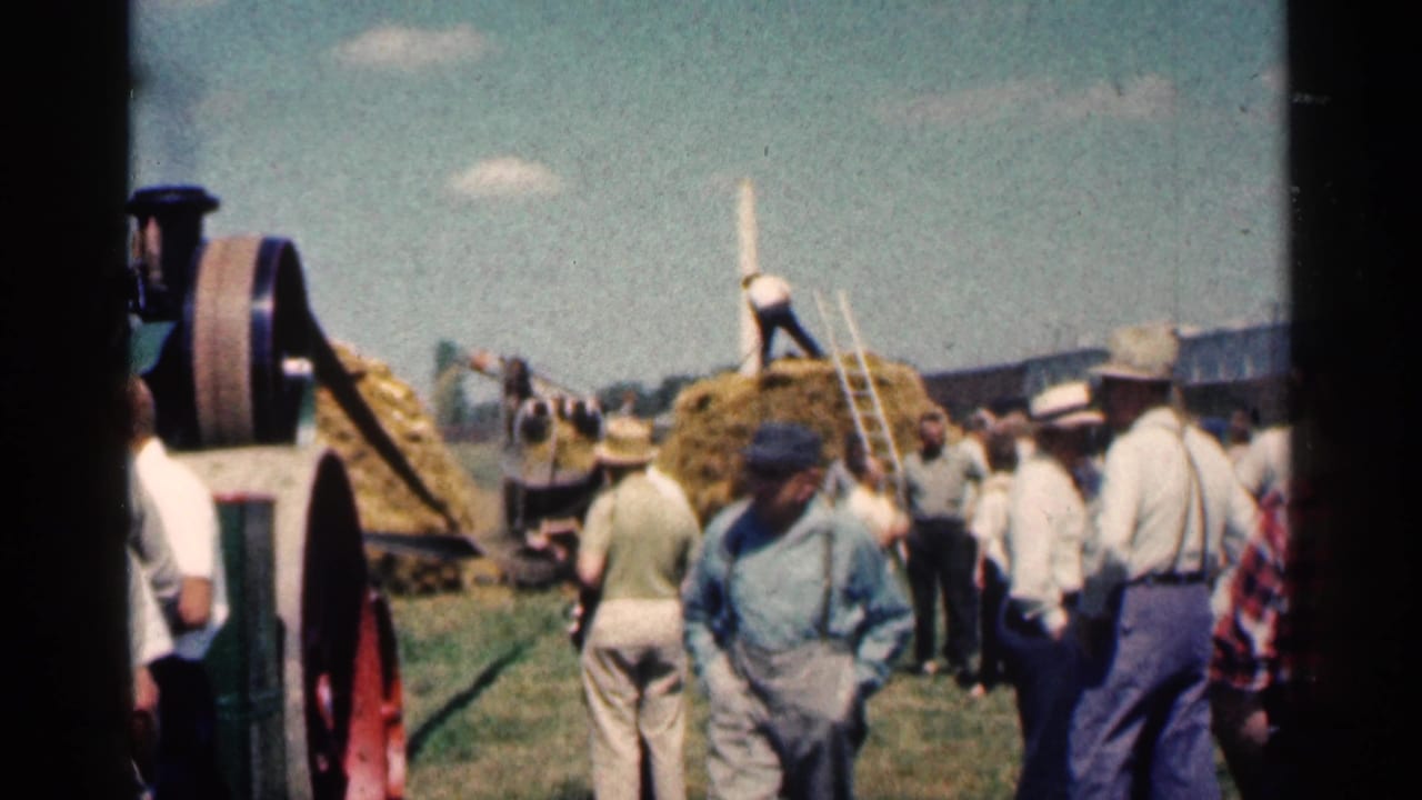People gather around hay bales and farm machinery in an outdoor rural scene.