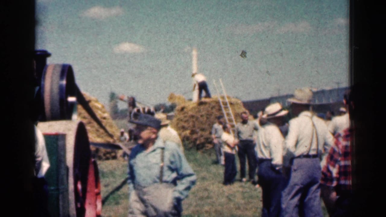 A man in work clothes walks past people watching hay stacking and farm machinery.