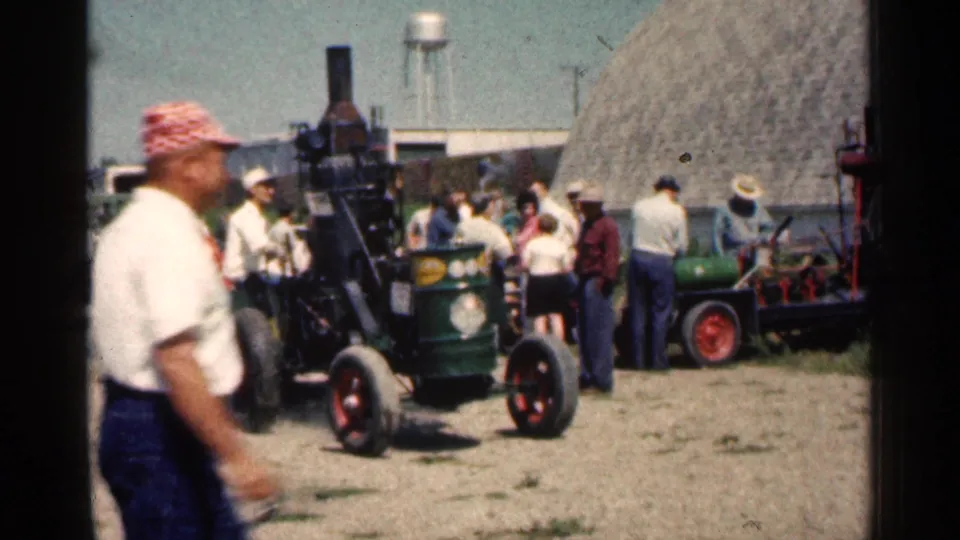 1963 Montgomery, Minnesota home-movie frame showing a small vintage tractor, spectators, a haystack, and a water tower in the background.