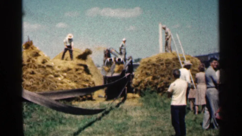 1963 Montgomery, Minnesota home-movie frame showing workers on hay piles, farm equipment, and spectators watching from the field.