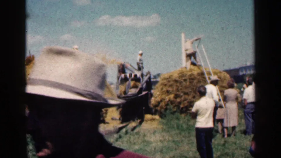 1963 Montgomery, Minnesota home-movie frame with a foreground hat partially blocking the hay harvest scene and workers in the distance.