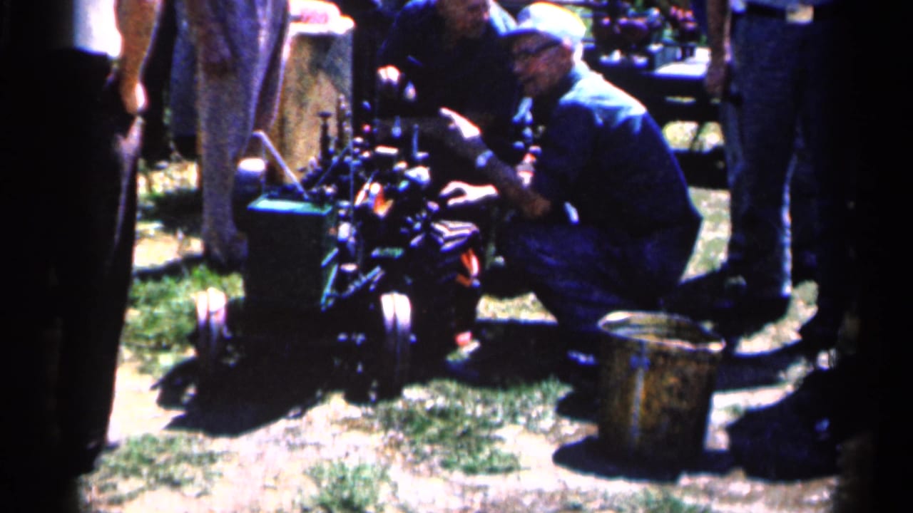 Men gather around a small engine on grass at an outdoor event.