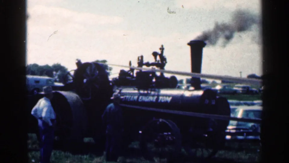 A large steam engine at the 1963 Montgomery, Minnesota outdoor event emits dark smoke while men stand near the machinery and cars sit beyond it.
