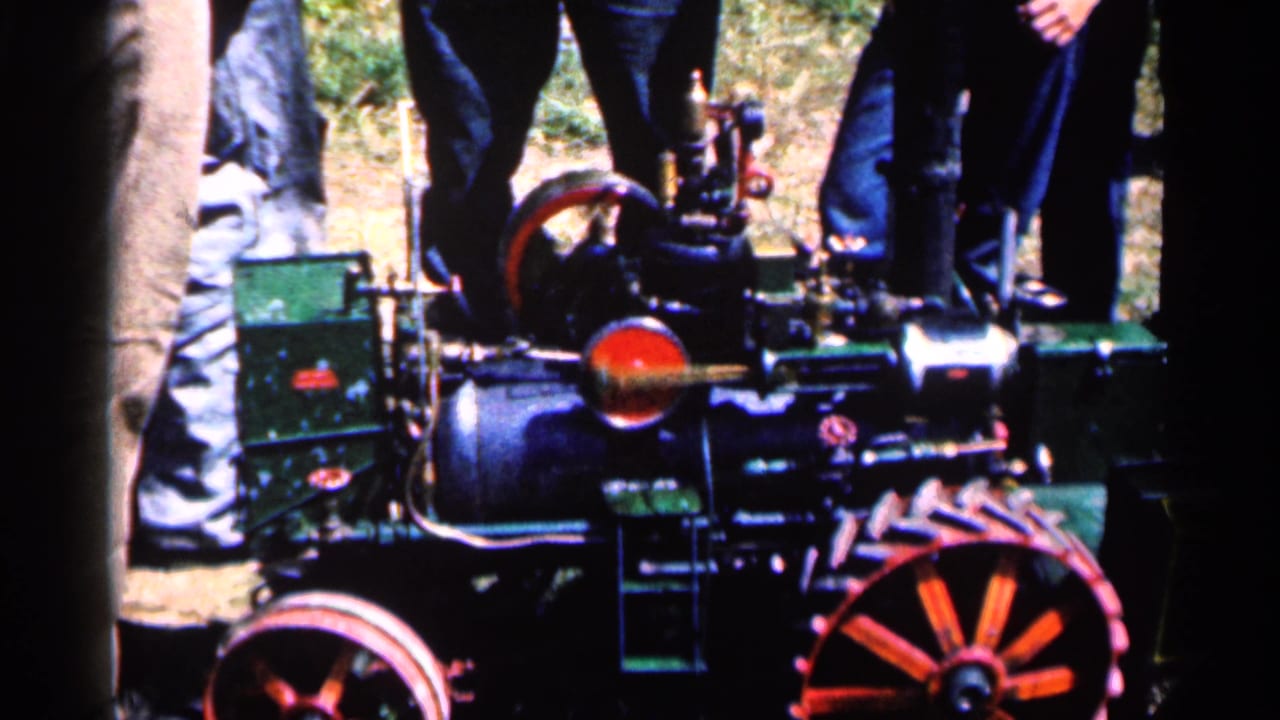 Close view of a small steam engine with red wheels and people standing behind it.