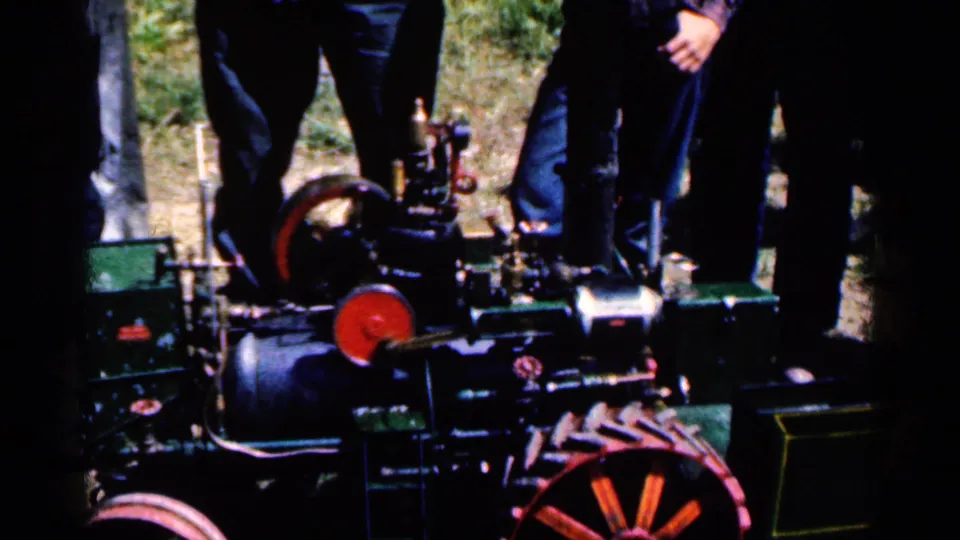 1963 home-movie frame showing the small steam engine from a low angle, with red flywheel details and onlookers gathered close around it.