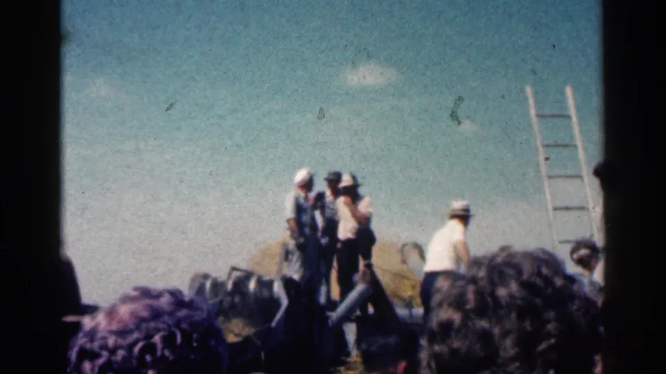 1963 Montgomery agricultural scene with workers on a raised platform or equipment beside a ladder, seen from a home-movie crowd viewpoint.