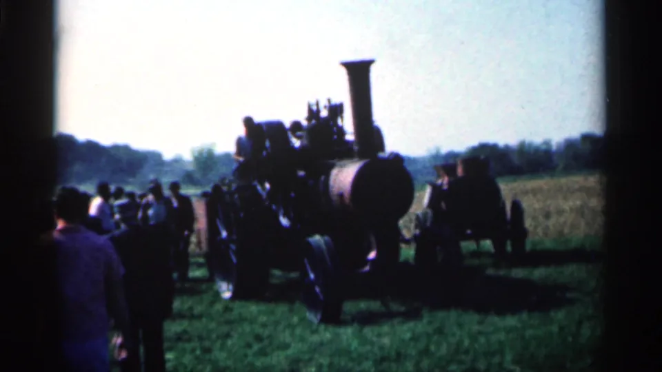 Men gather near a dark steam tractor during a 1963 Montgomery, Minnesota farm display, with open field and trees behind them.