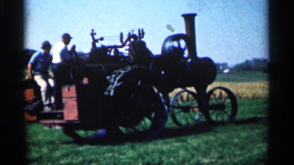 Two men appear to demonstrate or inspect the steam tractor from a side platform, with large spoked wheels visible in the field.