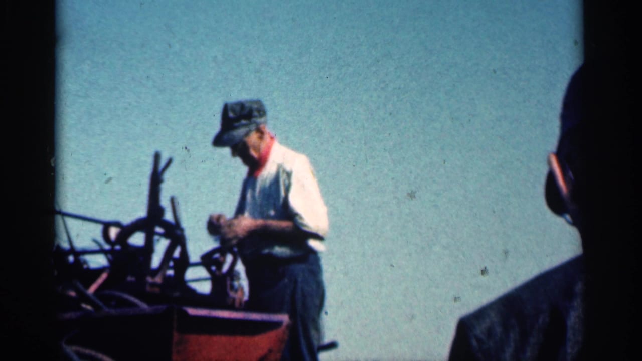 Man in cap and work clothes standing beside farm equipment under a bright sky.