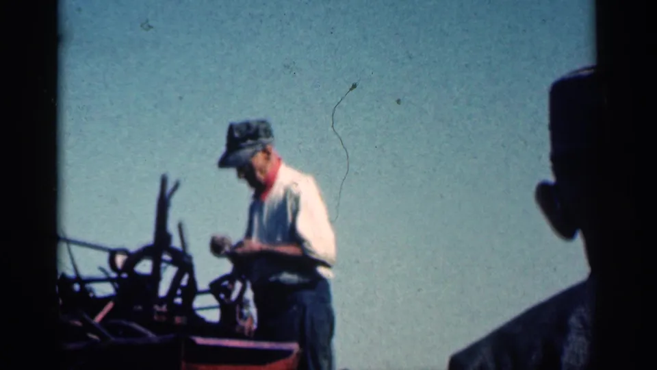 A man near farm machinery is seen in soft 8mm color, with a dark foreground figure or edge partly obscuring the frame.