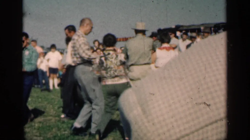 The home-movie camera looks across a busy outdoor dance, showing adults in 1960s clothing moving among a crowd on grass.
