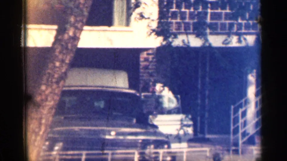 Soft 8mm frame showing a dark sedan parked outside a Texas brick building, with an adult near the open trunk.