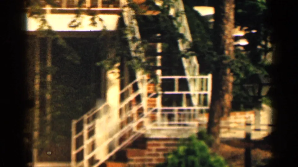 1963 Texas home-movie frame emphasizing white railings, brickwork, steps, foliage, and a partially visible person near the building.
