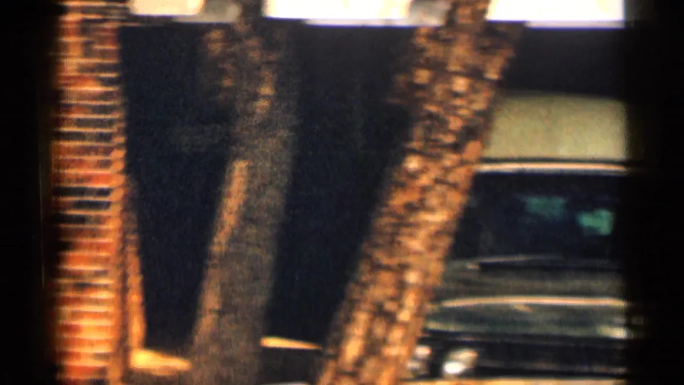 8mm frame from Texas in 1963 with tree trunks, brick facade, dark doorway, and the front portion of a parked sedan.
