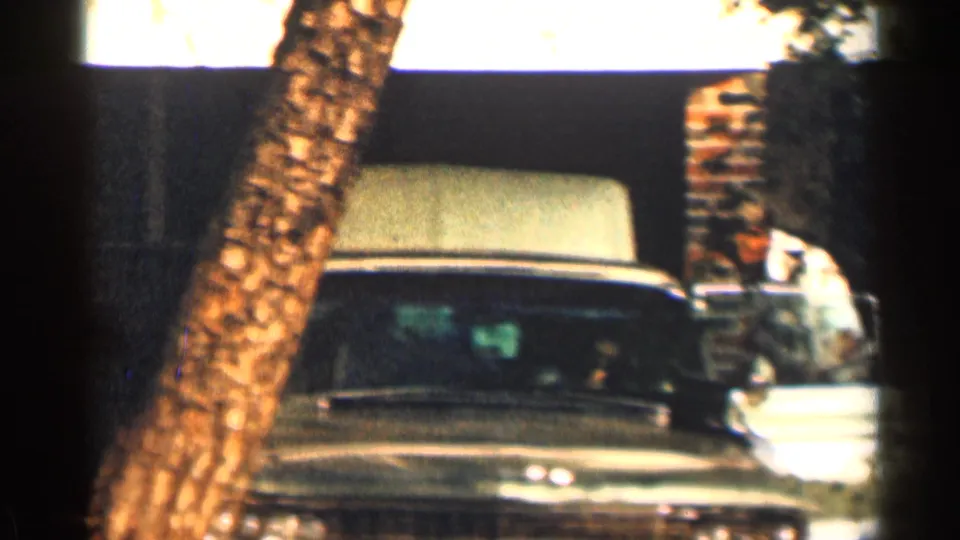 1963 Texas home-movie frame with an olive-green mid-century car parked by a tree beside brick residential walls.