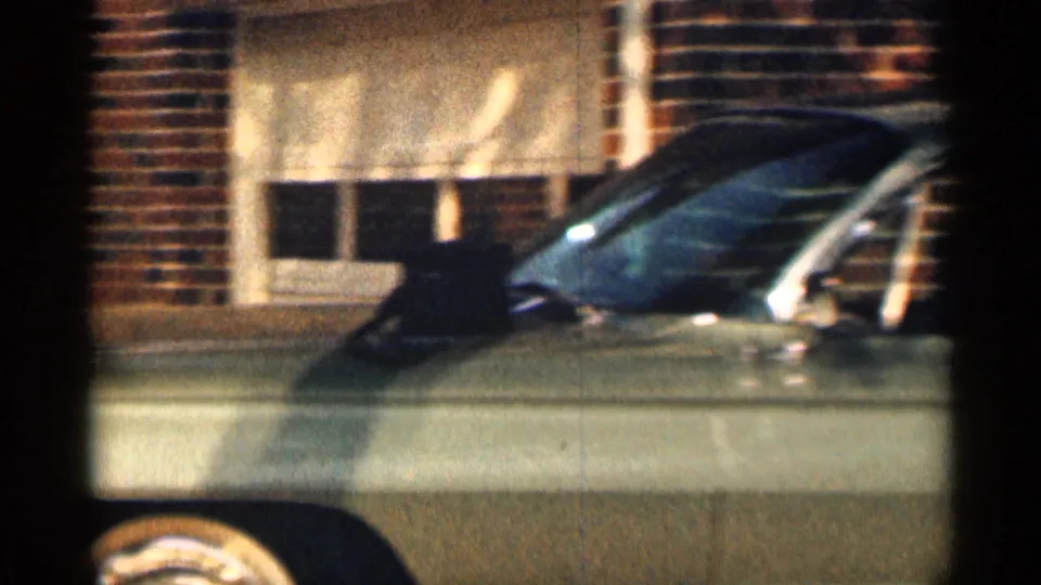 1963 Texas frame showing the side of an olive-green classic car parked in front of brick residential exteriors.
