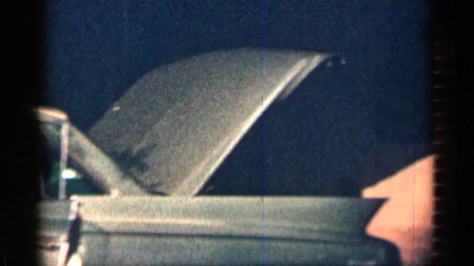 Close side view of the raised hood on a light-colored sedan in 1963 Texas home-movie footage.