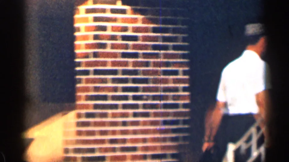 A man in a white shirt and dark pants walks away near brickwork and a stair rail in the Texas 1963 home-movie scene.