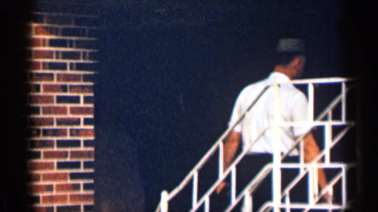 1963 Texas home-movie frame of an adult man walking up white metal stairs beside brick and dark exterior walls.