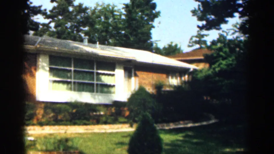1963 Texas home-movie view of a ranch-style brick house with broad windows, lawn, shrubs, and mature trees.