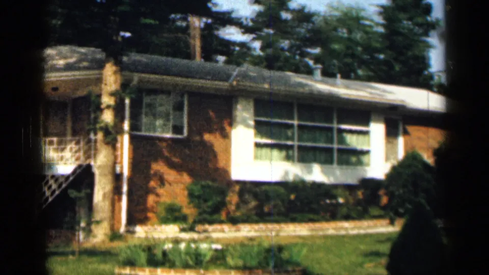 1963 Texas home-movie frame showing a brick ranch house, wide front windows, landscaped lawn, and partial exterior stairs.