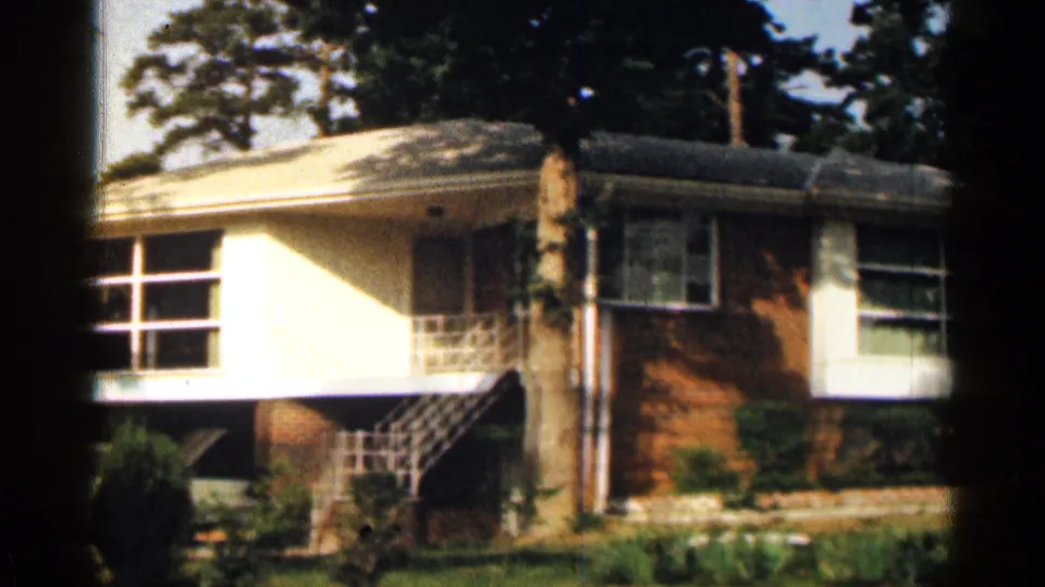 1963 Texas home-movie view of a brick ranch house with white railings, shaded porch area, windows, lawn, and trees.