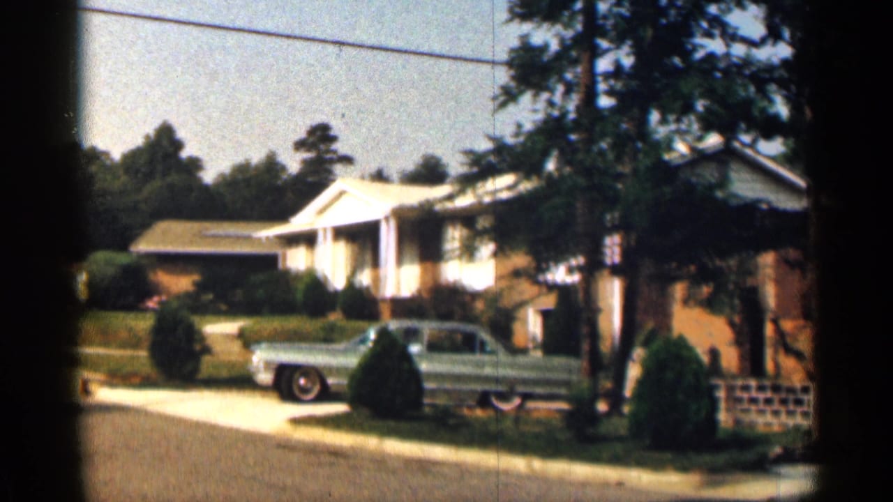 In a 1963 Texas residential street scene, a blue mid-century sedan sits near a driveway outside a brick ranch-style house with a white portico and trimmed shrubs.
