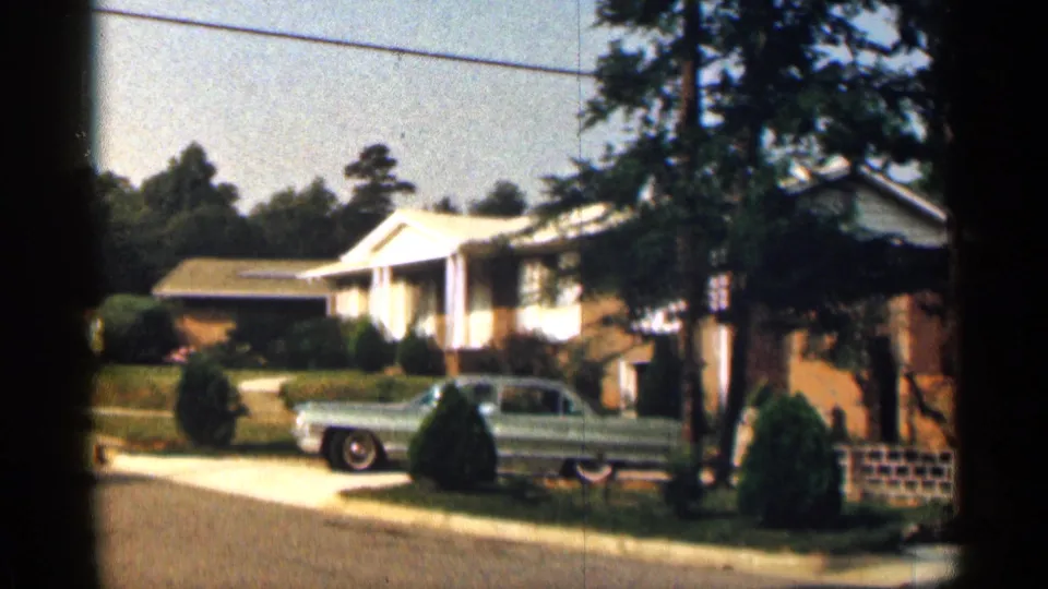 In a 1963 Texas residential street scene, a blue mid-century sedan sits near a driveway outside a brick ranch-style house with a white portico and trimmed shrubs.