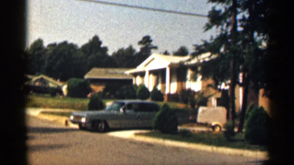 The blue sedan begins moving along the residential street while towing a small covered trailer past a brick house and manicured front lawn in Texas.