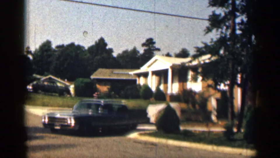 The sedan and trailer turn out from the driveway area onto the quiet suburban street, with a brick home, lawn, shrubs, and trees in the background.