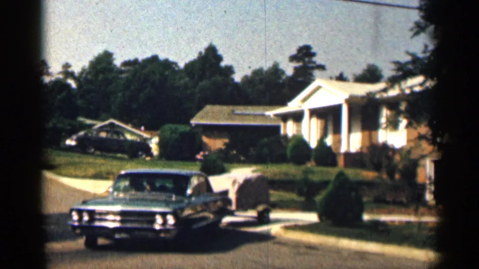 The blue sedan faces the camera as it pulls the small covered trailer down the residential street, with the ranch-style house and front garden still visible behind it.