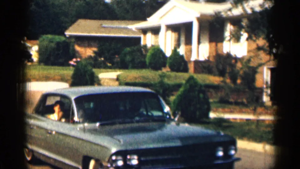 A 1963 Texas suburban street view shows a light-colored American sedan passing the camera with an occupant visible.