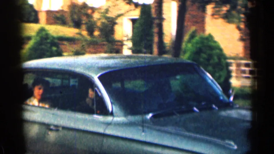 Close 8mm frame of a classic sedan passing in a Texas residential neighborhood, with adult occupants visible inside.