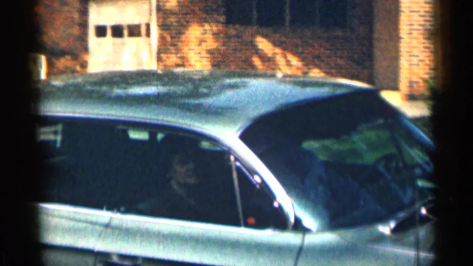 The sedan continues past a brick suburban home in 1963 Texas, with the car roof and side windows filling the frame.