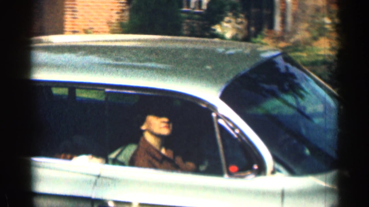 Woman seated in the passenger side of a classic car near brick buildings.