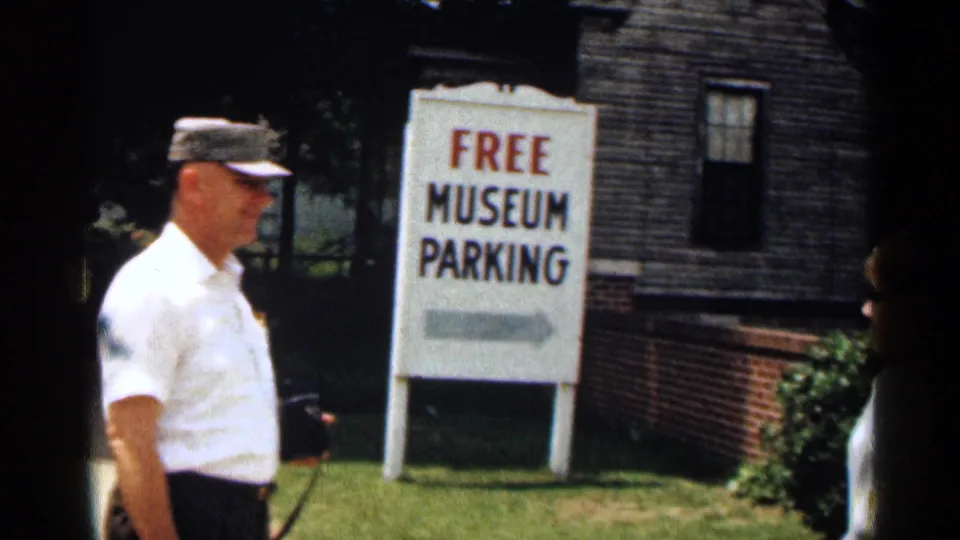 1963 Texas home-movie frame of a man in a cap near a FREE MUSEUM PARKING sign and brick exterior.