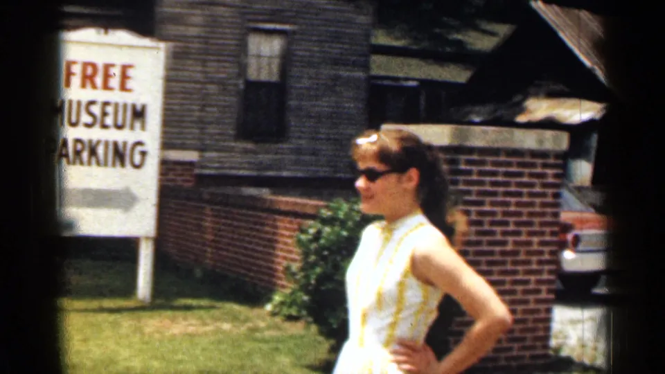 1963 Texas frame of a woman in sunglasses and a striped dress near a Free Museum Parking sign outside town buildings.