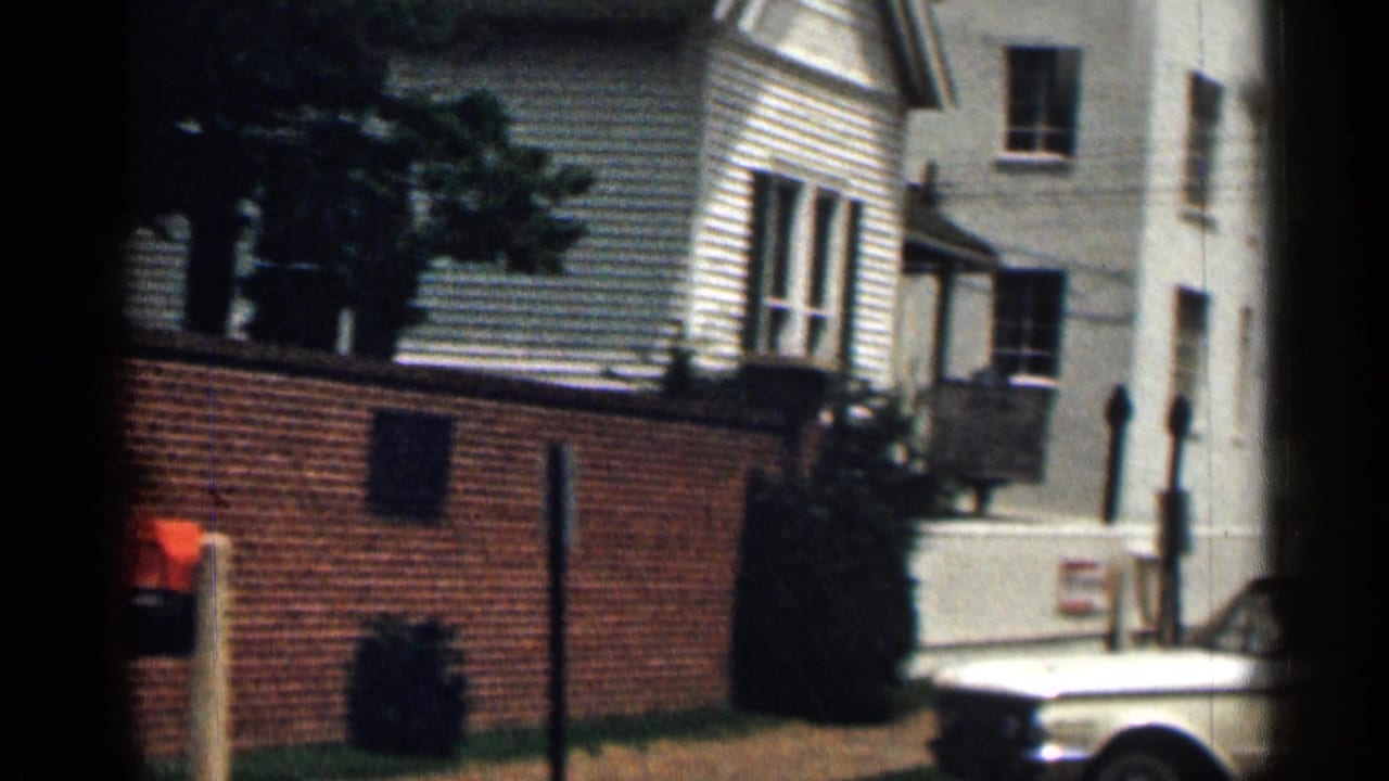 White older car parked beside a brick wall and light-colored houses.