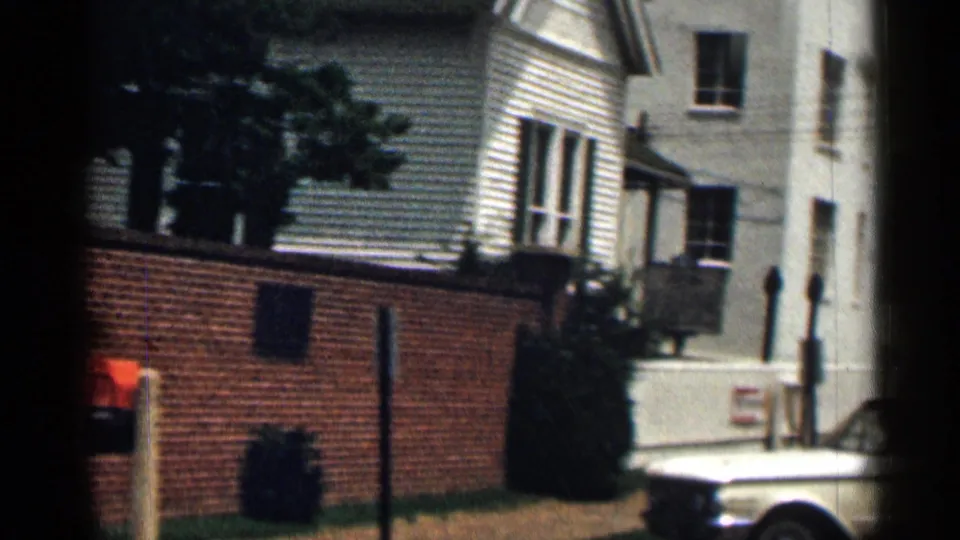 1963 Texas street view with a white older car near a brick wall, clapboard houses, shrubs, and sidewalk details.