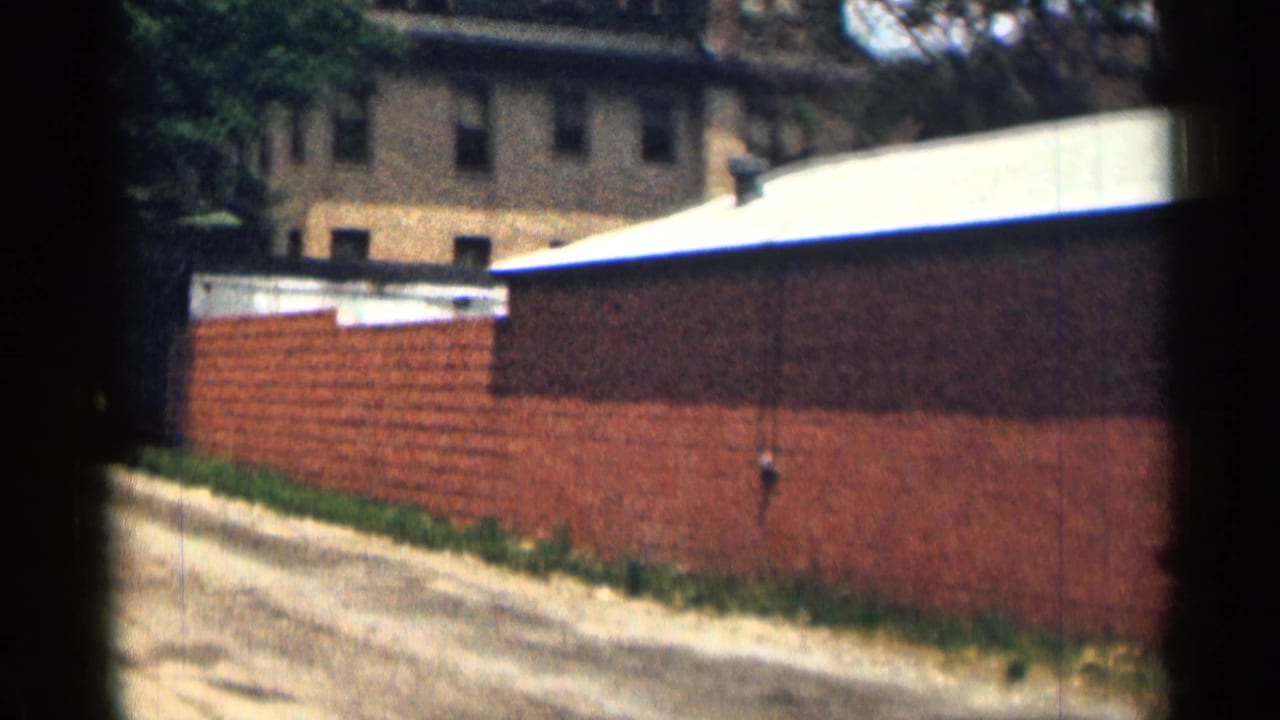1963 Texas 8mm frame showing a red brick wall along a dirt road, with a low brick building and trees in the background.