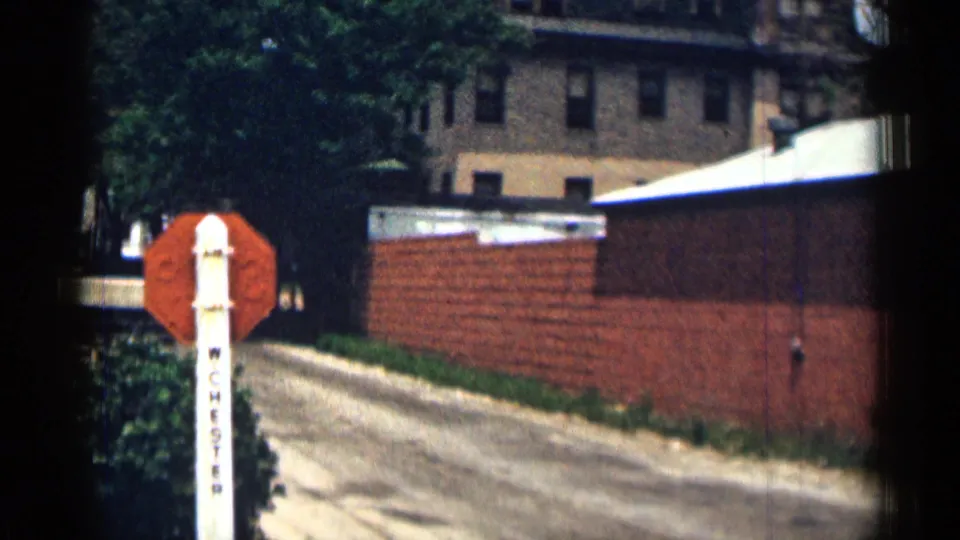 1963 Texas roadside view with a red stop sign, white W CHESTER post, brick wall, dirt road, and buildings beyond.