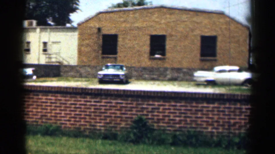 1963 Texas exterior with a foreground brick wall, parked mid-century cars, grass, and a tan brick building with dark windows.