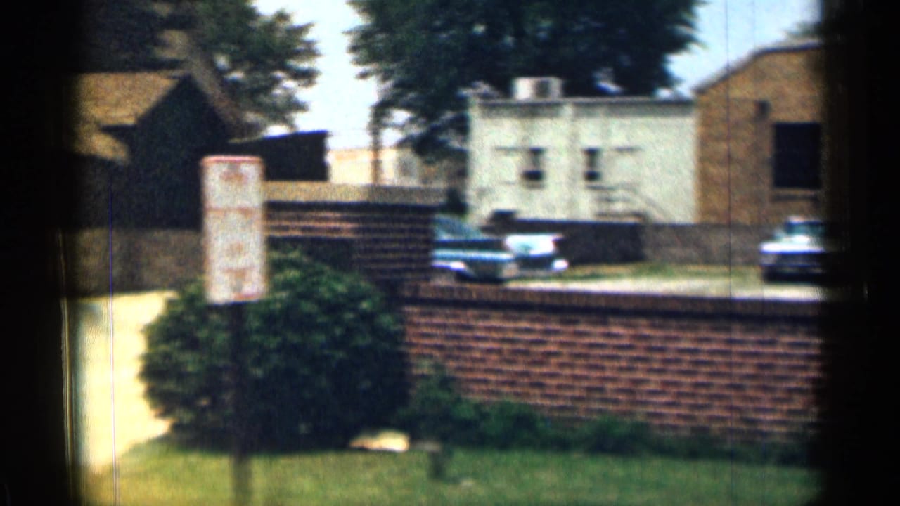1963 Texas 8mm frame showing a redbrick wall, pale building, parked cars, grass, shrubs, and a white museum parking sign.