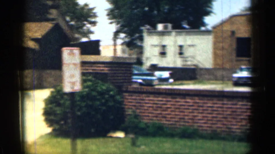 1963 Texas 8mm frame showing a redbrick wall, pale building, parked cars, grass, shrubs, and a white museum parking sign.