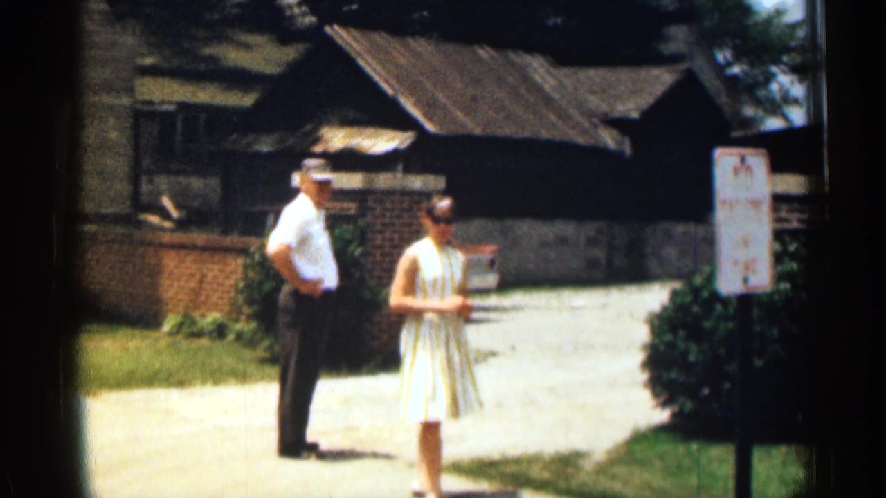 Man and woman standing near a redbrick wall and parking area outside low buildings.