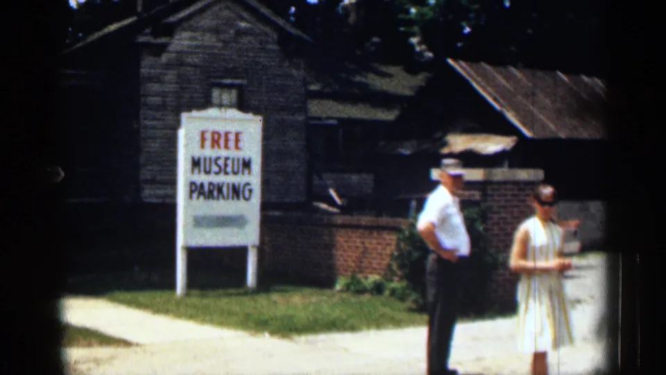 White FREE MUSEUM PARKING sign stands beside a path, with two adults, grass, redbrick wall, and dark wooden building visible.