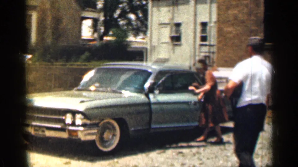 1963 Texas home-movie frame of a woman in a brown dress at a blue sedan while a man in a white shirt and cap stands beside the driveway.